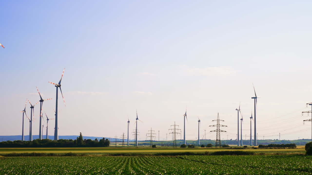Farmlands with silhouettes of wind turbines and electricity power supports. Clear blue sky at backdrop with a plane. Low angle view.