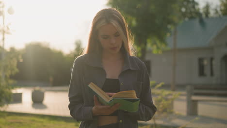 dama en ropa gris de pie al aire libre hojeando las páginas de un libro, con la luz del sol suave iluminando a su alrededor y un fondo borroso de árboles y un edificio
