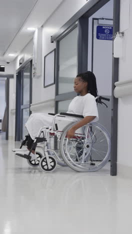 vertical video of african american female patient in wheelchair in hospital corridor, slow motion