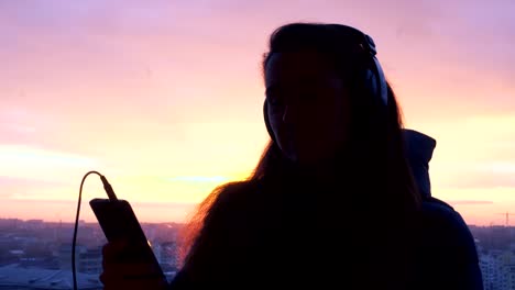 silueta de una mujer bailando y escuchando música en el teléfono en la ciudad al atardecer