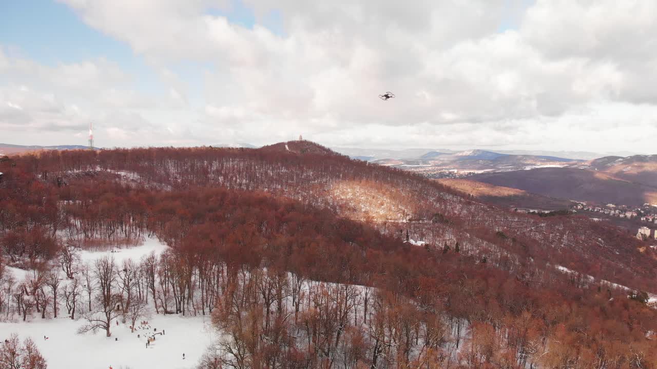 Aerial pan following a drone flying over a beautiful winter landscape with snowy hills, forest and a city in the background