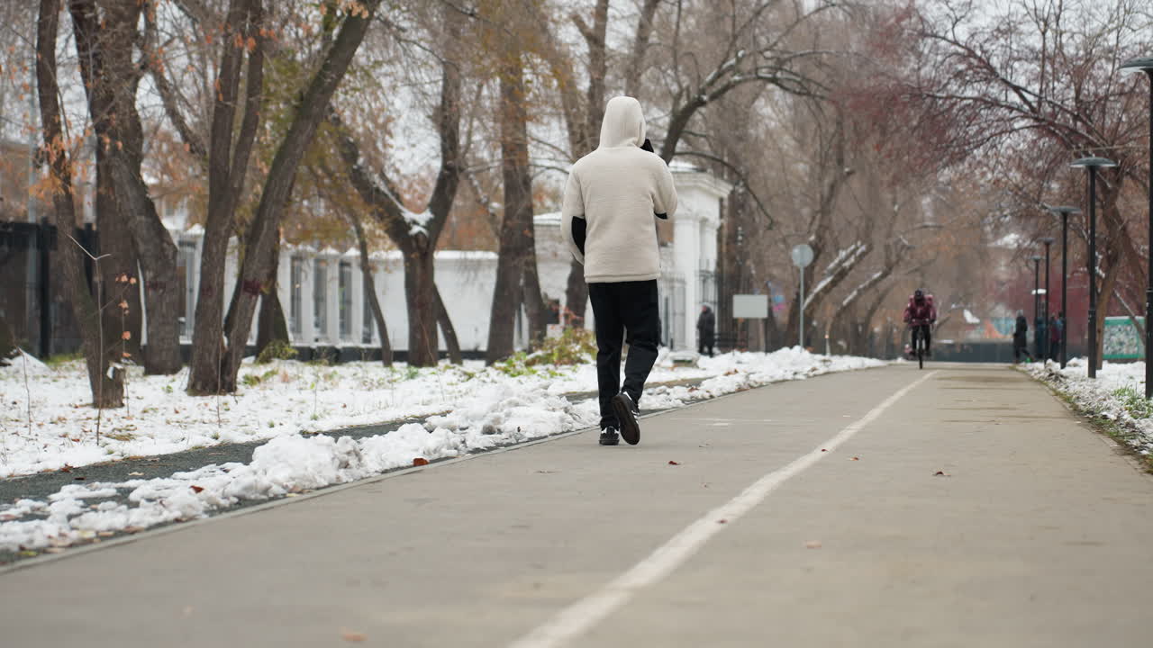 전화 통화 중인 젊은 남자의 뒷면은 겨울에 거리를 걸어다니며 자전거를 타고 다가오는 누군가의 흐릿한 모습, 바닥의 눈과 배경의 나무입니다.