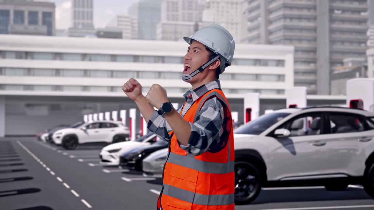 近代都市の充電ステーションで電気自動車が充電されている中、安全ヘルメットを着用したアジア人男性エンジニアが作業中に目標達成を叫び、喜びを表現している横向きの姿。