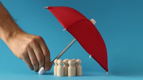 hand holding red umbrella over wooden figures, symbolizing protection and coverage