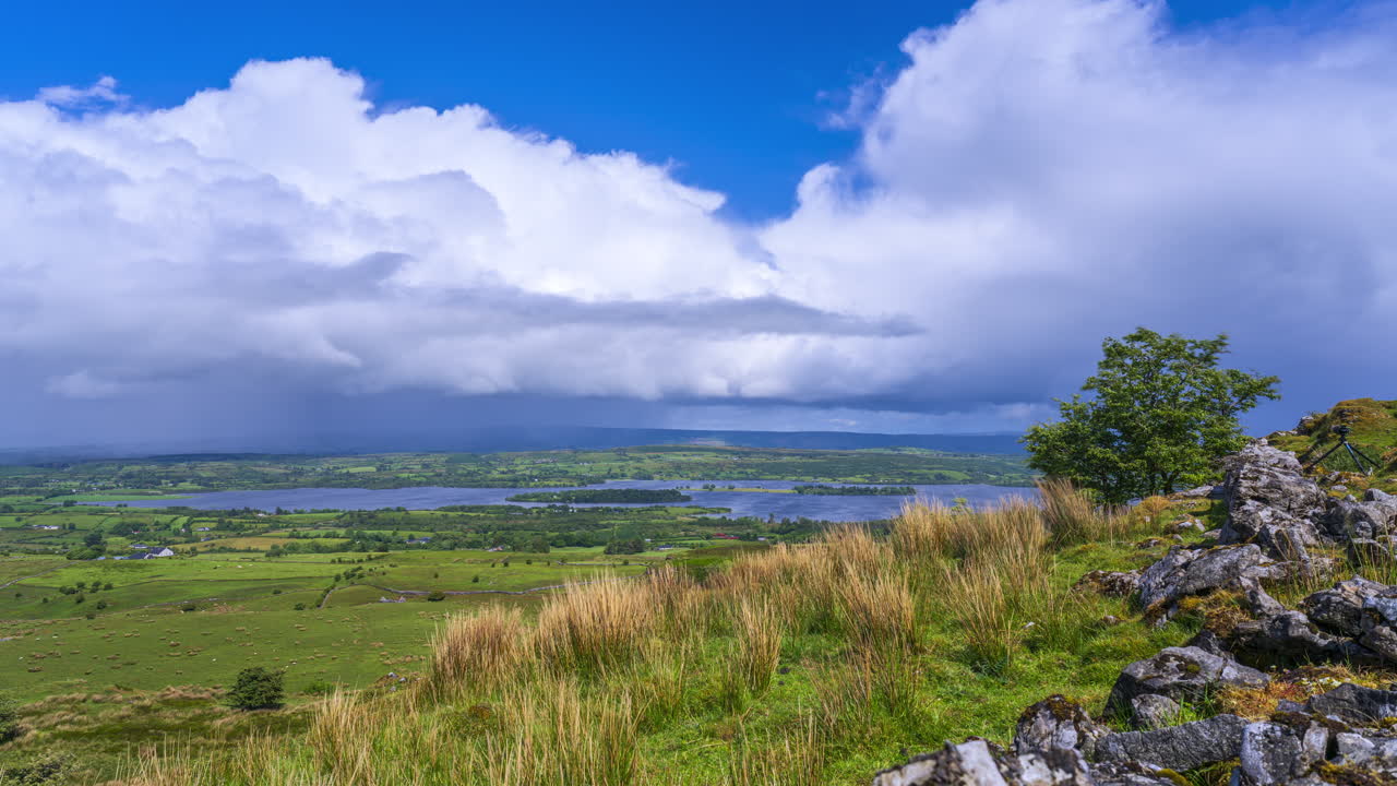 Tidsforløb af et enkelt træ i en landskabsdal med en forbigående regnstorm, bakker og en sø i det fjerne, på en forårsdag med skyer og sol, set fra Carrowkeel i Sligo County i Irland.