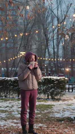 joven con una servilleta en la mano y comiendo maíz mientras está de pie en el parque con nieve, luces de vacaciones en el fondo, momento relajado al aire libre, disfrute de la comida