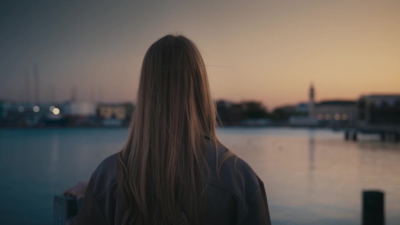 A close-up of a woman standing by the water at Valencia Marina during sunset, with a clock tower and waterfront buildings silhouetted against the warm sky in the background.
