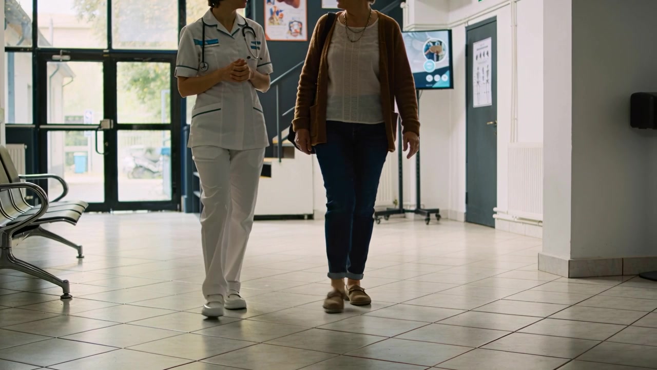 Enfermeira de uniforme e idoso discutindo cuidados, caminhando da porta no saguão, ela carregando bolsa e estetoscópio.