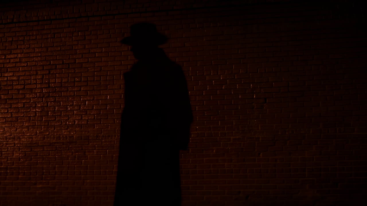 Ombre sur un mur de briques la nuit, silhouette d'une personne en fedora et trench-coat, éclairage cinématographique tamisé.