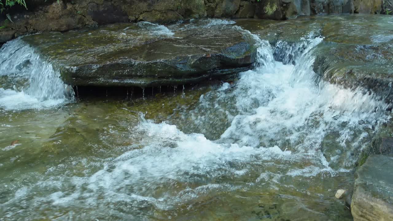 Acqua limpida di un ruscello che scorre impetuosa su rocce piatte in una piccola cascata, catturando il movimento naturale, il flusso fresco e un tranquillo paesaggio all'aperto in un ambiente roccioso di torrente.