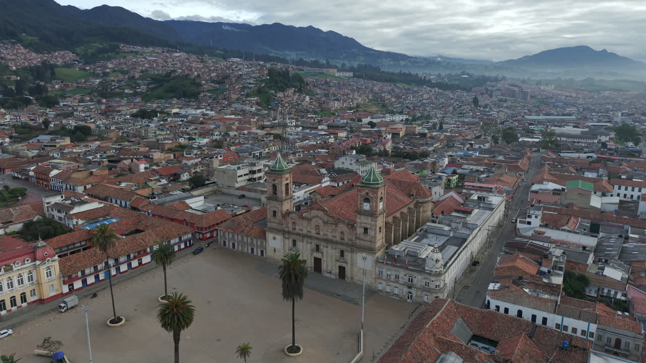 Drone footage showcases La Catedral de Zipaquirá's stunning architecture, nestled amid a vibrant townscape under a moody sky, with expansive mountainous horizons