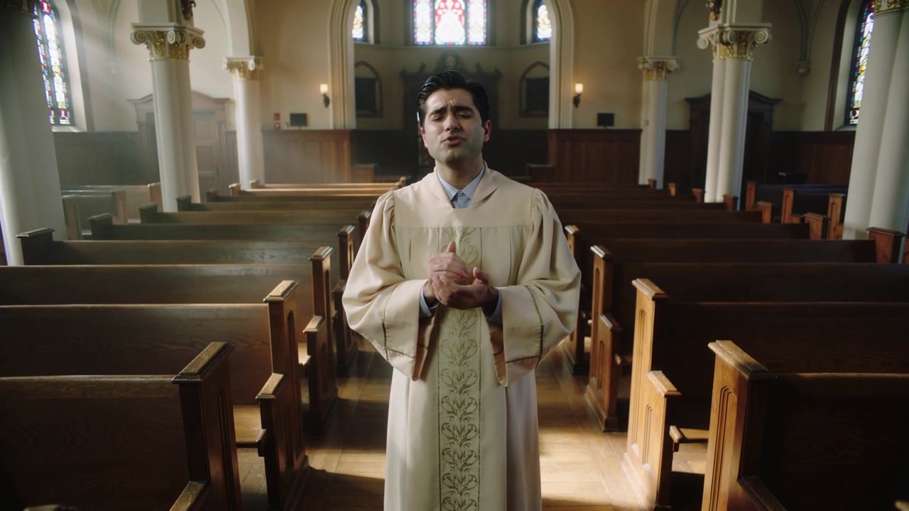 Prêtre officiant levant les mains, serrant sa poitrine dans la nef, un hymne commence, en chasuble, lunettes de soleil