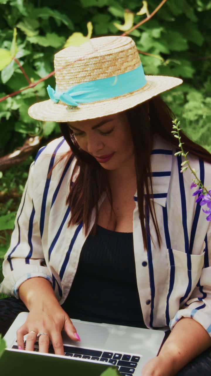Young woman with a straw hat engaged in online work while surrounded by greenery, showcasing a relaxed and focused atmosphere in a natural setting