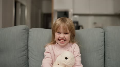 pretty happy little girl in casual wearing sitting on sofa with toy dog and smiling