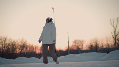 swaying girl around under sunset light in snowy park wearing puffer coat beanie and headphones enjoying playful winter movement and joyful energy amid bare trees and pastel sky reflecting serene vibe
