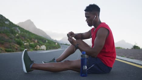 african american man wearing earphones using smartphone while sitting on the road