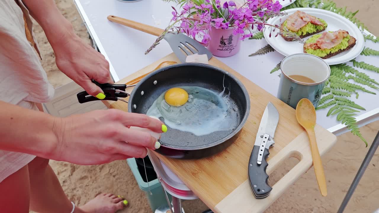 Cracked egg sizzling in pan on rustic camping table with flowers food and coffee