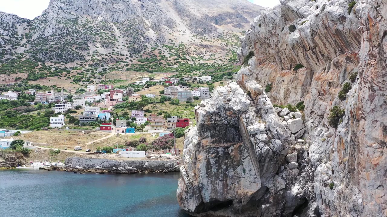 Kuzey Fas'ın manzaralı kıyı görünümü; sakin Akdeniz sularının üzerinde yükselen devasa kayalık uçurumları ve arka planda engebeli dağ yamaçlarının altına gizlenmiş küçük bir köyü gösteriyor.