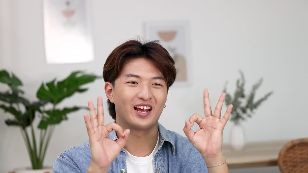 ชายชาวเอเชียยิ้มแย้มสวมเสื้อเชิ้ตยีนส์ ทำท่าโอเคสองมือขณะหัวเราะอยู่ในอาคาร