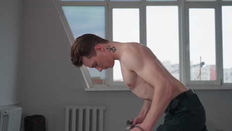 shirtless student with tattoos on neck and chest engaged in resistance rope workout in bright room with wide window, focused on body building and strength training amid natural light