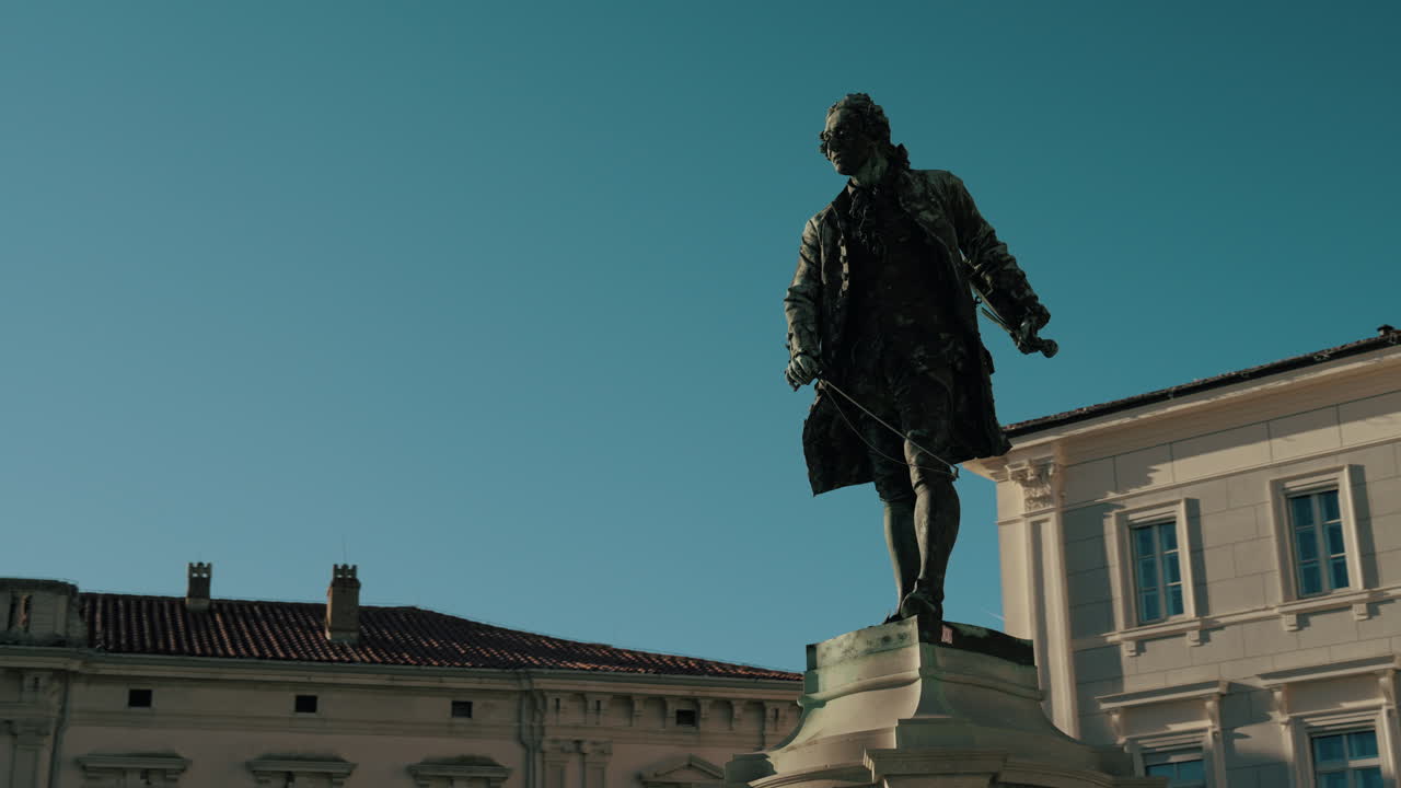 Statua di Giuseppe Tartini in Piazza Tartini, Pirano, Slovenia, con il municipio visibile sullo sfondo. Il filmato mostra la piazza storica sotto un cielo soleggiato e l'architettura circostante.