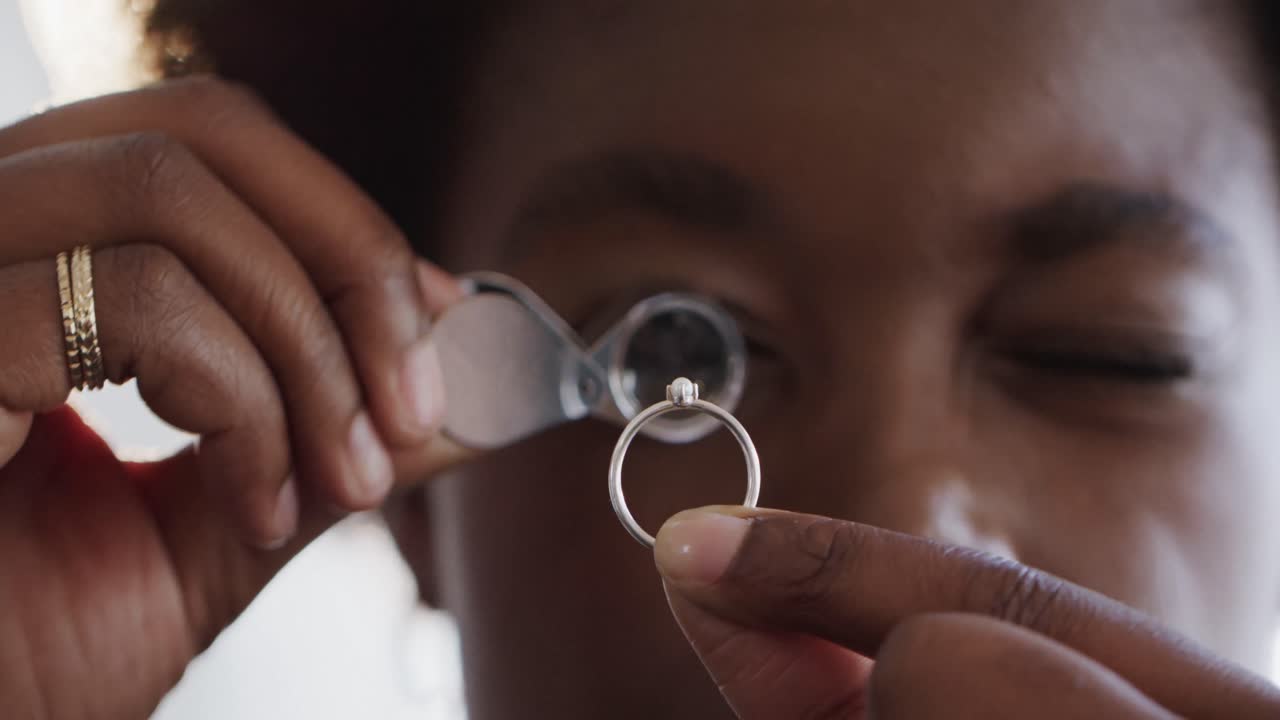 Afroamerikansk kvindelig arbejder inspicerer en ring med forstørrelsesglas på værksted i slow motion.