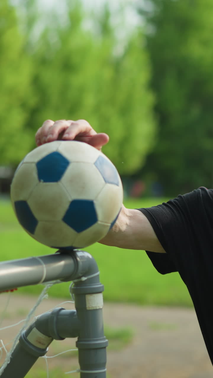 An elderly man in a black top rests his hand on a soccer ball placed on a goalpost, he stands calmly with, with a blurred view of a paved road and lush green grass