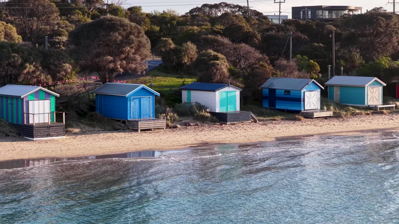 Drone pans over vibrant beach huts on sandy shore, calm water, soft daylight, tranquil mood