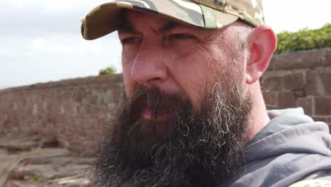 Bearded urban man wearing baseball cap with thoughtful expression looking across windy beach