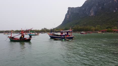 Drone-Viaja-Hacia-Adelante-A-Los-Coloridos-Barcos-De-Pesca-Asiáticos-En-La-Bahía-De-Khao-Ta-Mong-Lai-Con-La-Montaña-Rocosa-En-El-Fondo