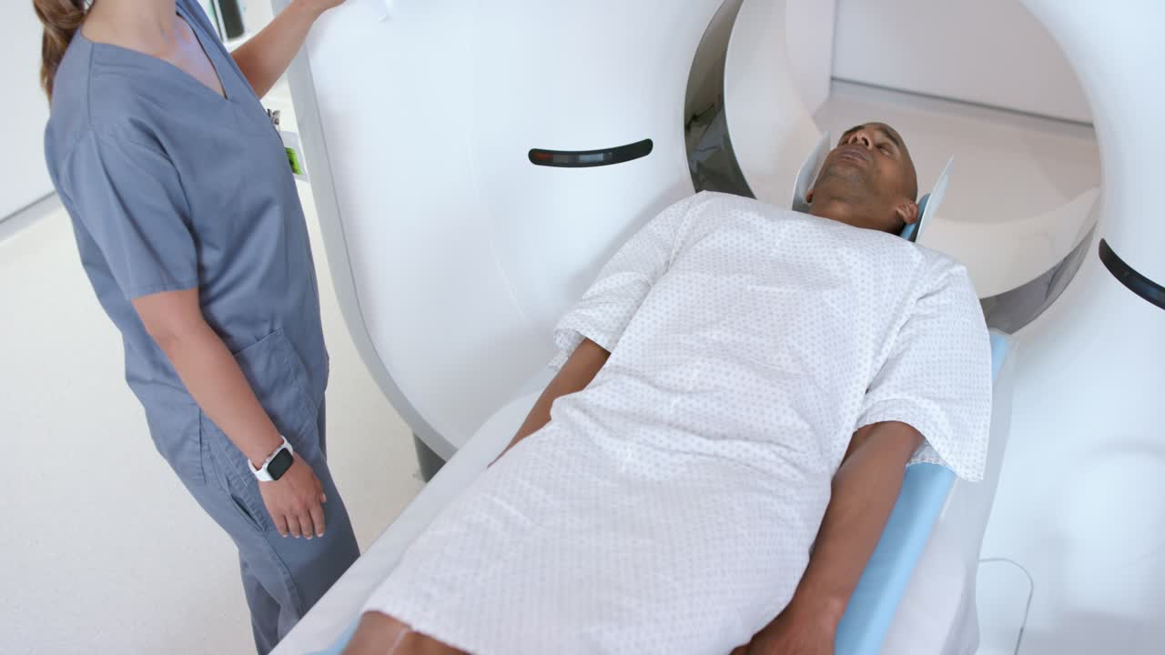 Tecnica afroamericana in camice che tocca il gantry e regola il tavolo, mentre guida il paziente maschio nella TC.