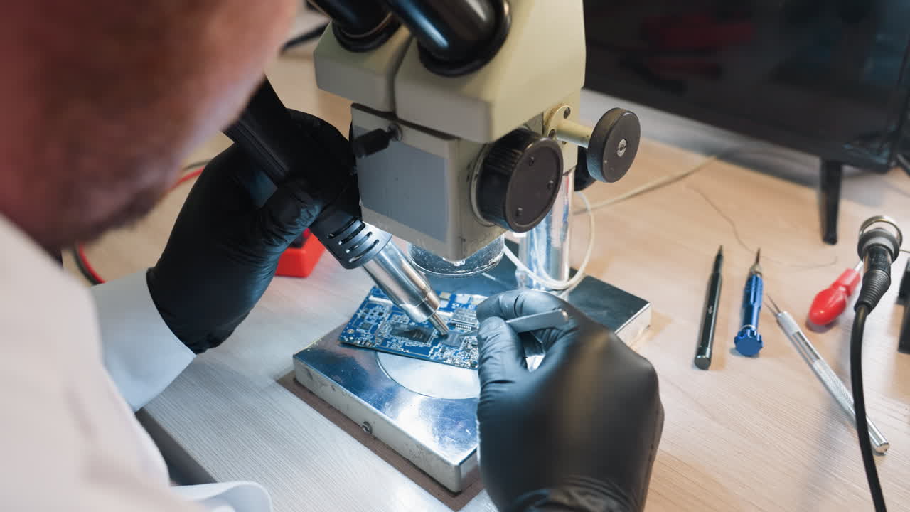 Close-up view of a technician in a lab coat using a microscope and soldering tools to meticulously repair a circuit board