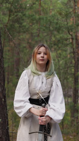 medieval princess in white dress sticks sword into ground in green forest. ancient clothes style and weapon. movie filming set natural decoration