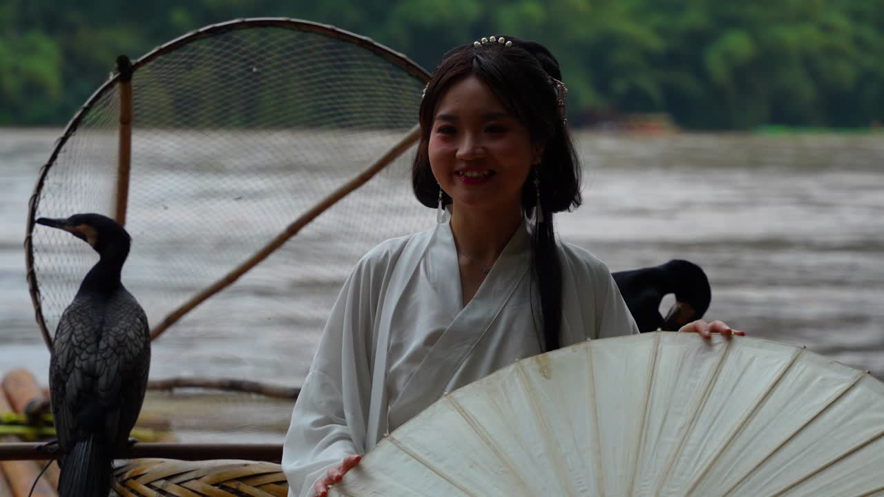 mujer sonriente vestida de hanfu y de pie en una balsa de bambú con pájaros cormoranes en el río li, china
