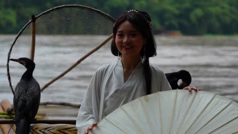 Mujer-Sonriente-Vestida-Con-Hanfu-Y-De-Pie-Sobre-Una-Balsa-De-Bambú-Con-Pájaros-Cormoranes-En-El-Río-Li,-China