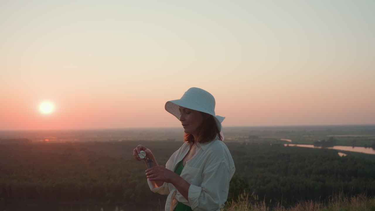 Young woman in white sun hat and light shirt gently blows bubbles while enjoying peaceful sunset view over lush green landscape and distant river