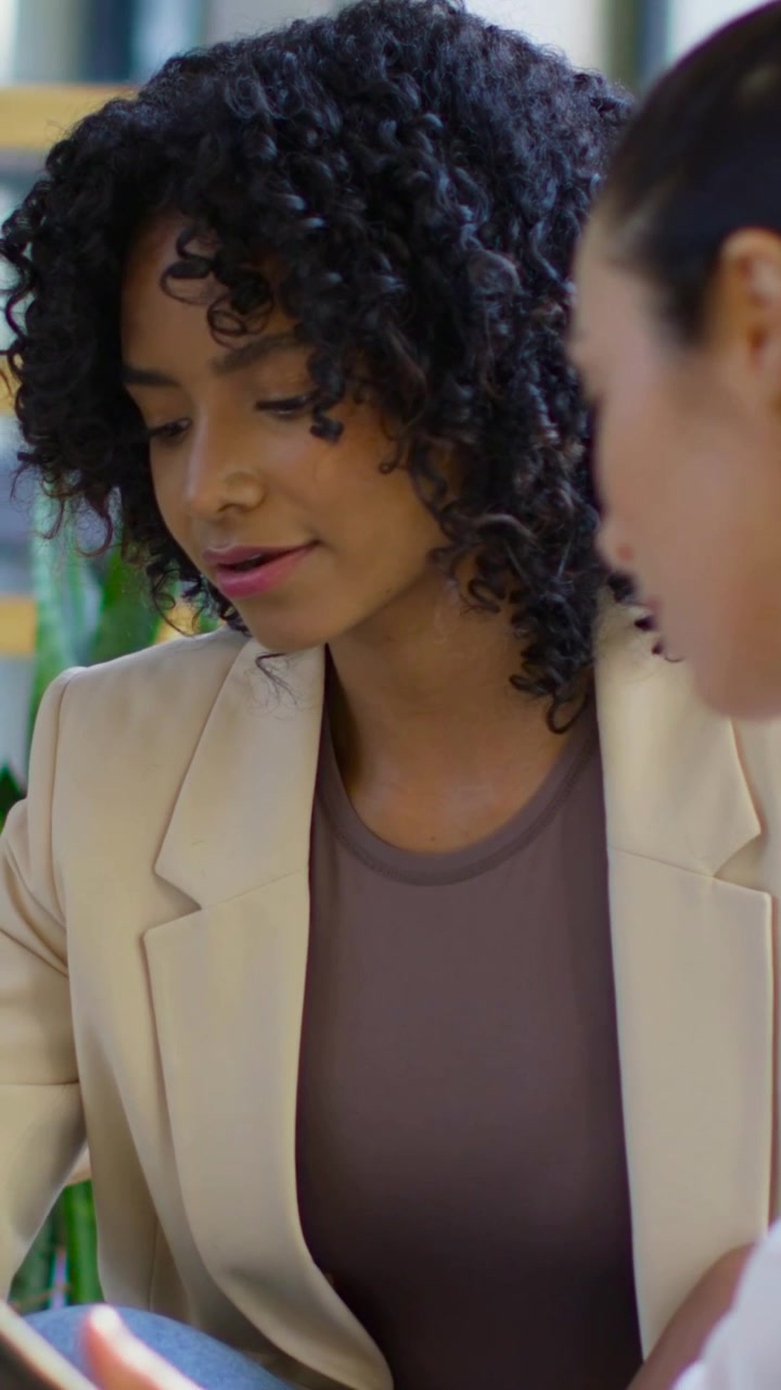 Vídeo vertical: Mulher em blazer bege sendo observada, colega de escritório entrando pela direita com tablet para revisão.