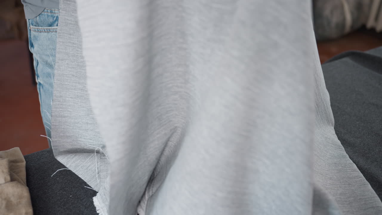 Costumiers arranging garments as one spreads out ash colored fabric on workspace table, carefully preparing textiles for design project in tailoring studio filled with sewing patterns and tools