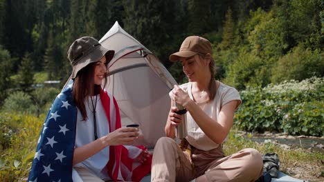 A-blonde-girl-in-a-cap-pours-tea-for-a-brunette-girl-who-is-wrapped-in-the-flag-of-the-United-States-of-America-near-the-tent-against-the-backdrop-of-a-green-forest