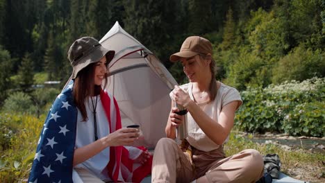 a blonde girl in a cap pours tea for a brunette girl who is wrapped in the flag of the united states of america near the tent against the backdrop of a green forest