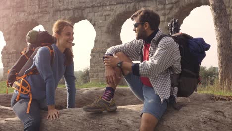 happy young couple backpackers tourists sitting on a log trunk talking and smiling in front of ancient roman aqueduct ruins in parco degli acquedotti park in rome at sunrise with guitar sleeping bag slow motion steadycam