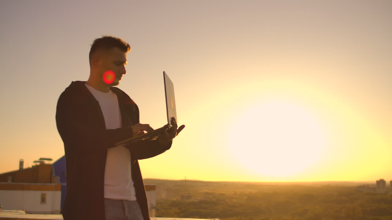 Hacker che usa il laptop sul tetto con vista sulla città e grafico forex. Concetto di hacking e statistiche. Un uomo al tramonto al rallentatore che scrive codice software su un laptop