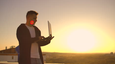 usando un portátil en el techo con vista a la ciudad y gráfico de divisas. concepto de hacking y estadísticas. un hombre al atardecer en cámara lenta escribiendo código de software en un portátil hacker