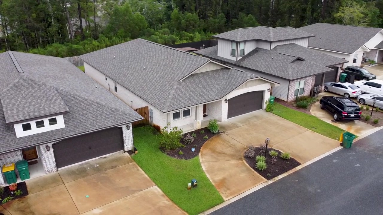 Vista aérea de drone de casas suburbanas com jardins frontais paisagísticos, garagens e entradas de veículos ao longo de uma rua residencial, mostrando o layout organizado do bairro