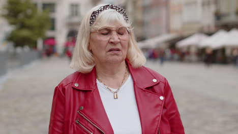 an older woman with blonde hair and glasses looks away from the camera while standing on a city street.