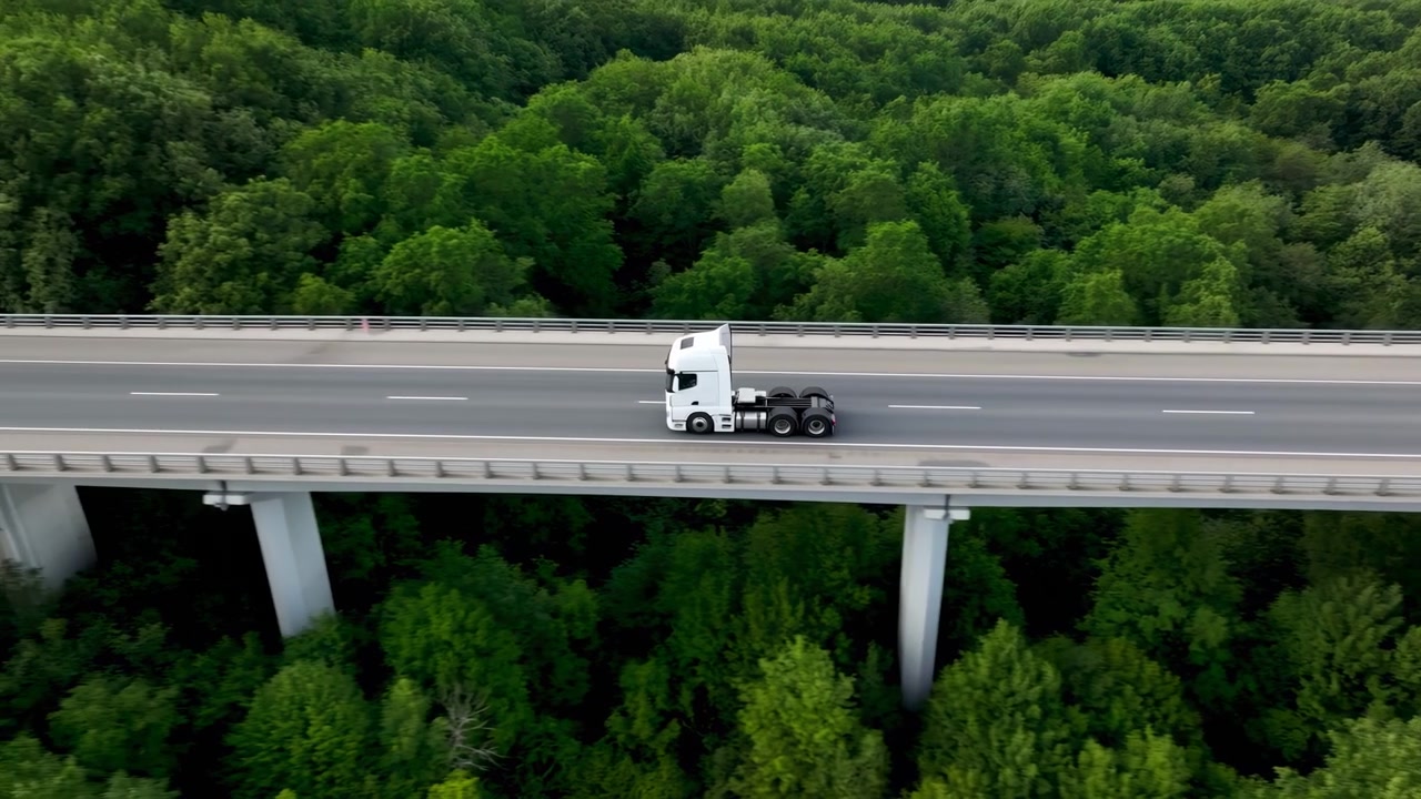 Un singolo trattore stradale bianco senza rimorchio che attraversa un ponte autostradale in cemento sopraelevato sopra una fitta chioma di foresta decidua continua, visto da un'angolazione aerea obliqua elevata con cielo coperto