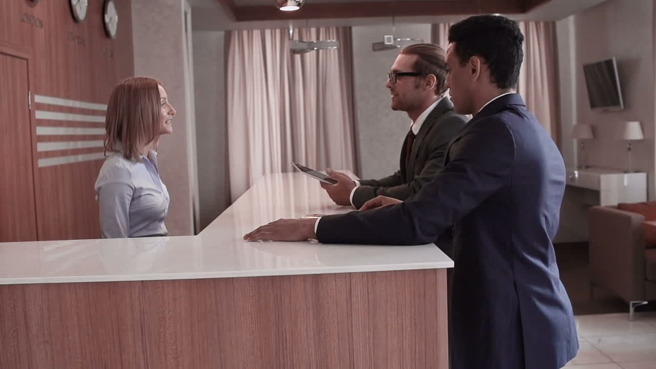 два бизнесмена у стойки регистрации отеля в аэропорту, один из них держит планшет и разговаривает с администратором