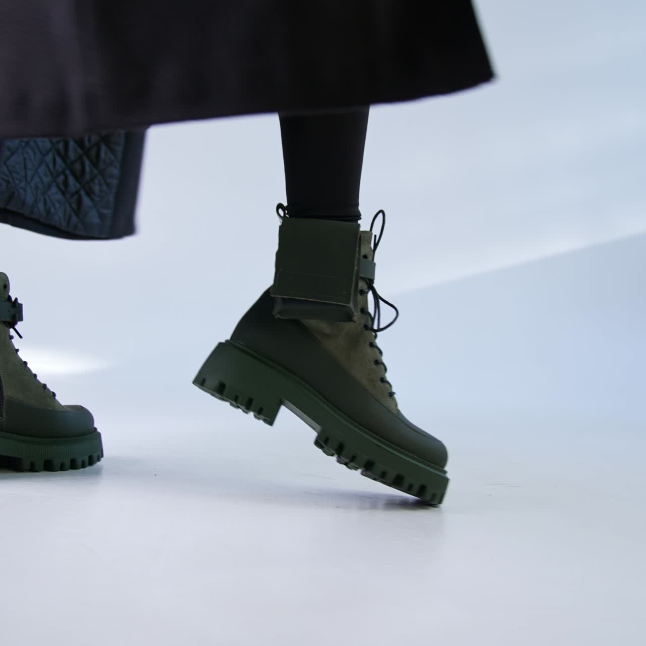 Woman wearing khaki combat ankle boots. Model in black clothes and long coat has fashionable footwear on showing it in studio