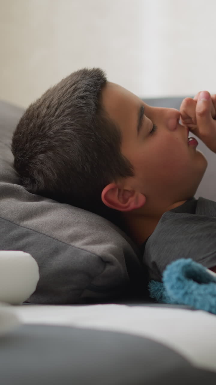 Niño enfermo acomodándose en el sofá, tapado con una manta, con un pañuelo cerca de la cabeza, intentando sentirse cómodo mientras descansa, incómodo y mal en casa, recuperándose de una enfermedad