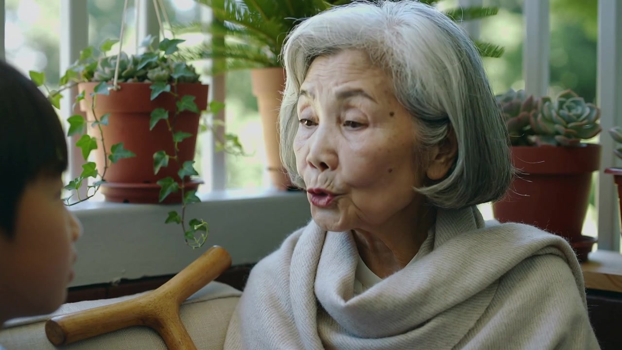 被男孩的目光所觸動，一位披著披肩的年長女士在陽光普照的窗邊說話並倚靠著，旁邊有手杖和植物。